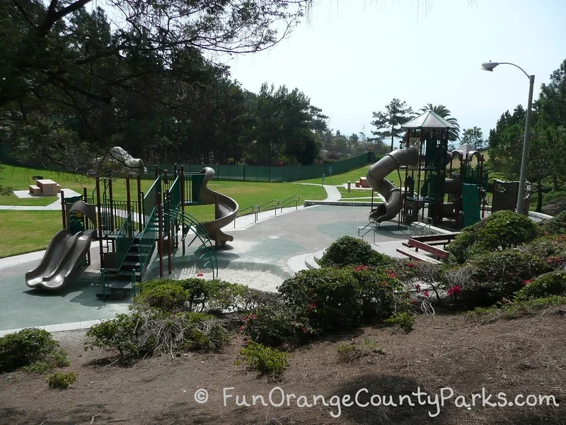 Pines Park playground with 2 play structures visible on recycled rubber pad with picnic tables and a grassy area