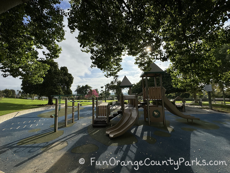 knotty pine park children's playground with slides on recycled rubber surface under the shade of trees with sun shining through partial clouds