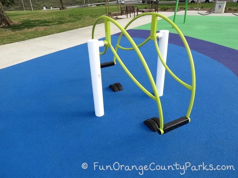 Standing teeter totter equipment at Pavion Park in Mission Viejo. Bright green bars on a blue recycled rubber surface.