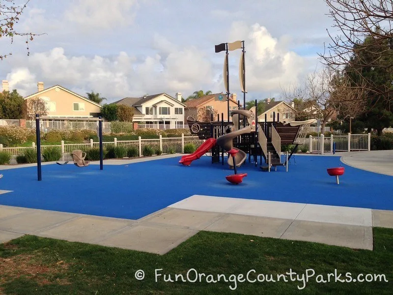 Ocean Breeze Park in Laguna Niguel with  red spinners and a pirate ship playground on a blue recycled rubber play surface. Swingset with chair swing and bench swing also visible. 
