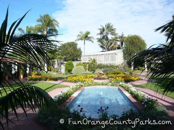 oblong blue water fountain with ornamental flowers and palms surrounding a white greenhouse building at Sherman Library and Gardens in Corona Del Mar