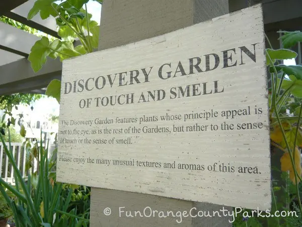 a worn white wooden sign with black text reading: "Discovery Garden of Touch and Smell. The Discovery Garden features plants whose principle appeal is not to the eye, as is the rest of the Gardens, but rather to the sense of touch or sense of smell. Please enjoy the many unusual textures and aromas of the this area."