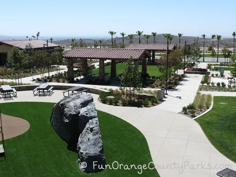 aerial view of picnic area at Baker Ranch Community Park with two shelters that have red tile roofs and a lawn area between them - a big climbing boulder sits below on the playground