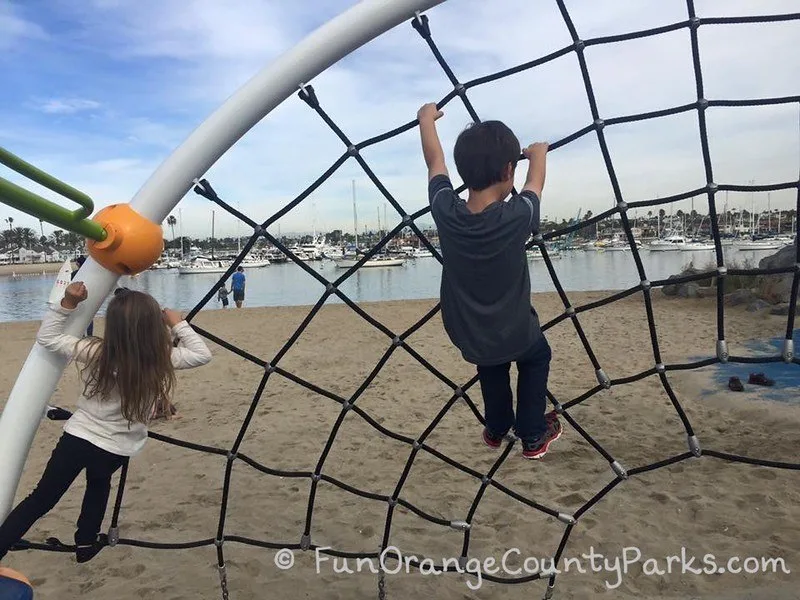 little girl and little boy on spiderweb play structure with sand and harbor view