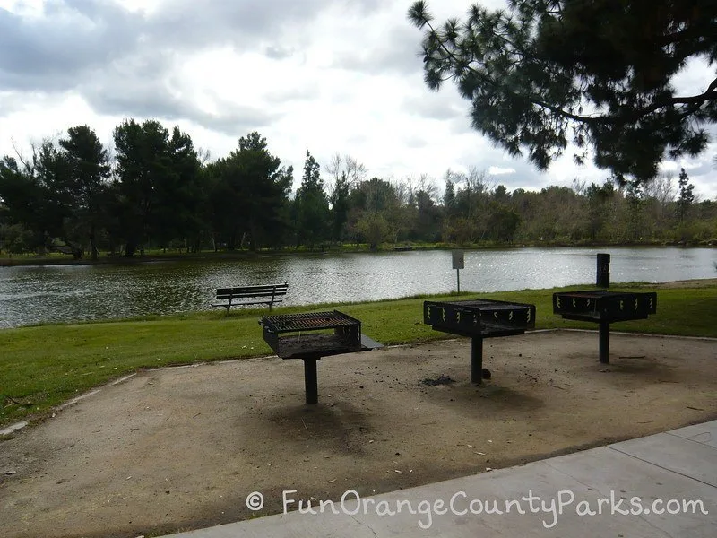 three large barbecue grills and a bench lakeside at Yorba Regional Park