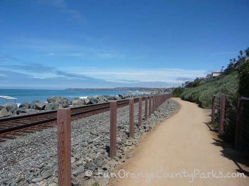 San Clemente Beach Trail with its dirt path down the center of the photo with the ocean and train tracks on the left with a green hillside on the right. A blue sky day with Dana Point Harbor visible in the distance.