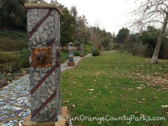 Five columns designed with mosaics of rock and tile along the Oso Creek Trail in Mission Viejo with a grassy area and sycamore tree.