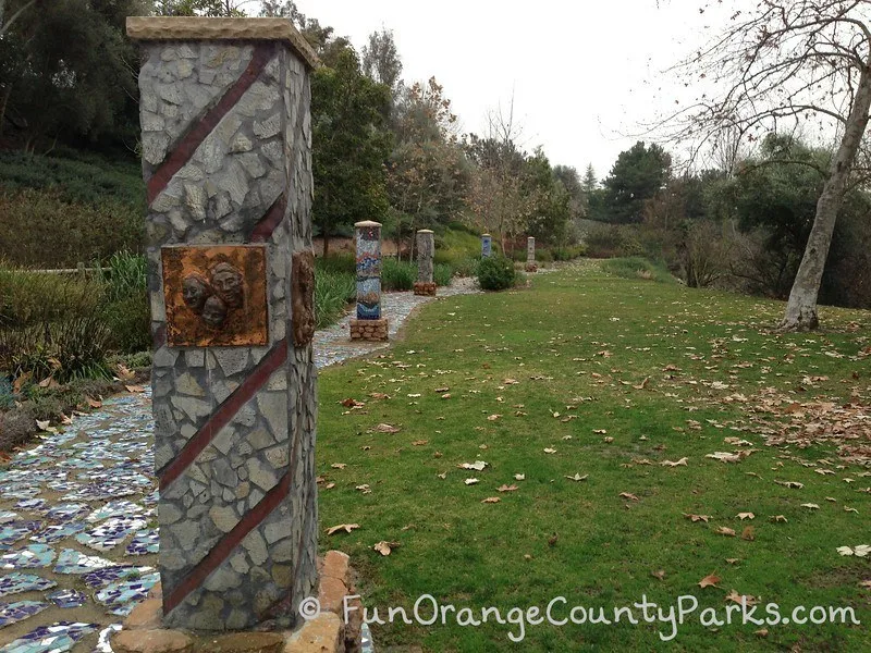 Five columns designed with mosaics of rock and tile along the Oso Creek Trail in Mission Viejo with a grassy area and sycamore tree.