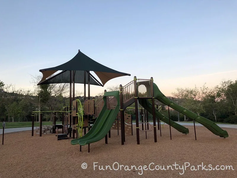 brown and green play structure on bark play surface at sunset