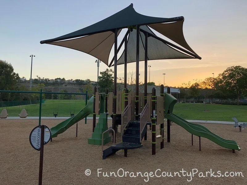 brown and green play structure with shade sails and a grassy area in the background