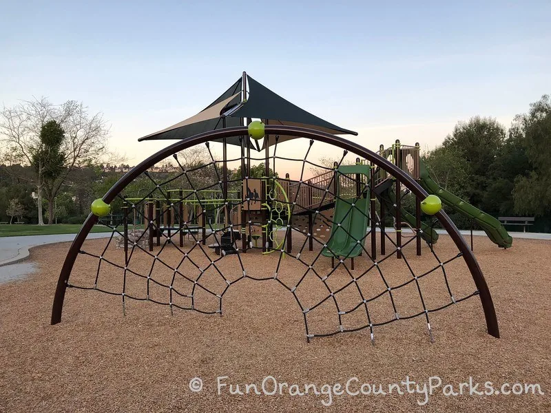 half circle arch on a bark play surface with black rope netting for climbing - playground in back