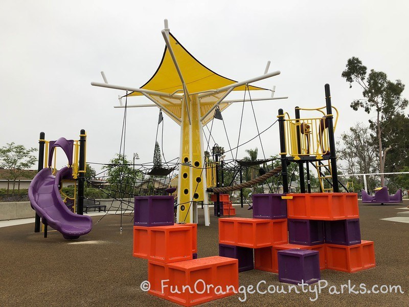 purple, orange, and yellow playground structures with blocks to climb, a short slide, a thick rope bridge and yellow shade sail at Cordova Park in Mission Viejo