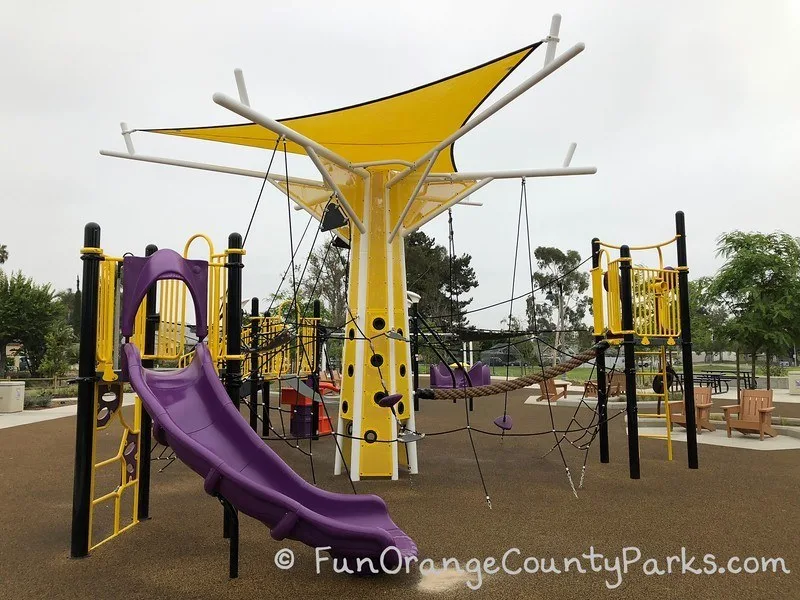 purple slide and views of the black rope netting and climbing opportunities on a brown recycled rubber surface that runs throughout Cordova Park playground