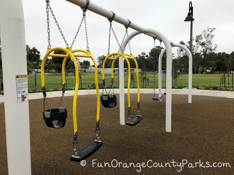 2 mommy and me swings with 2 bench swings above brown recycled rubber surface with grass field and school buildings behind the swings