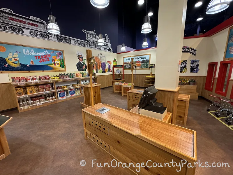 wooden grocery checker station with grocery store that looks like Trader Joes with food boxes on low shelves and murals on the walls