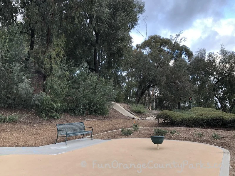 view of park with bench and bucket spinner on recycled rubber surface with trees in background
