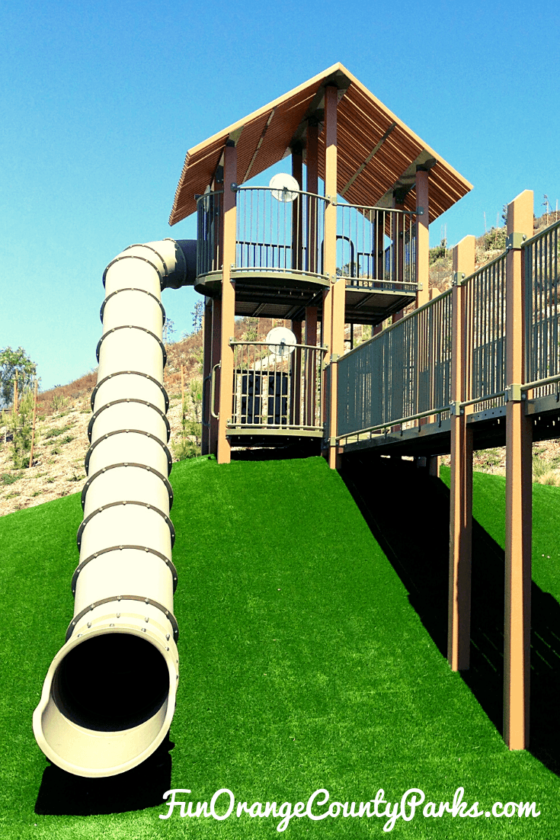 green astroturf hill topped with what looks like a treehouse playground and an enormous beige tunnel slide coming off the tower