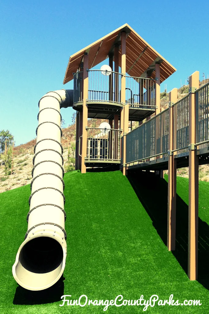 green astroturf hill topped with what looks like a treehouse playground and an enormous beige tunnel slide coming off the tower