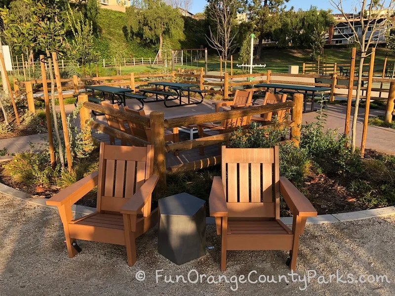 3 green metal picnic tables inside a fenced picnic area with two adirondack chairs and a side table for lounging - lawn area and play equipment in the background.