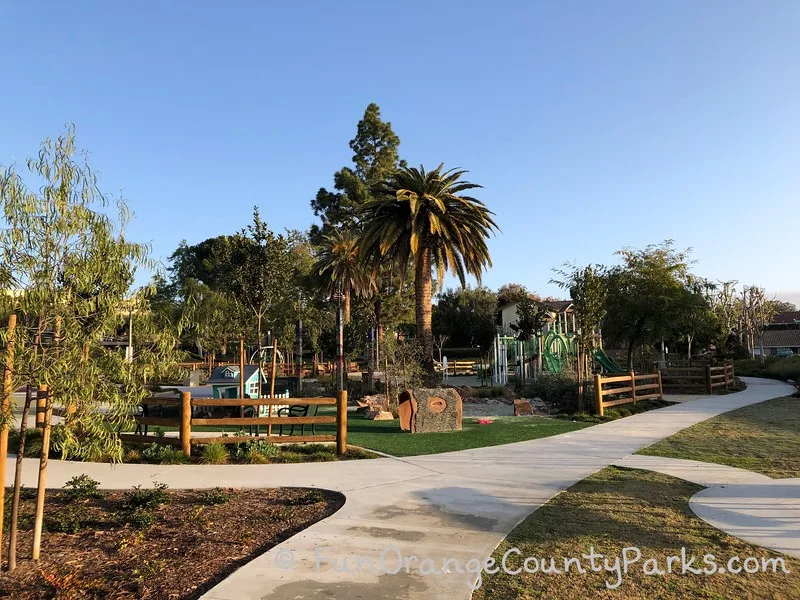 sidewalk into christopher park in mission viejo with big palm trees in the center of the playground. small play house and playground visible through the trees.