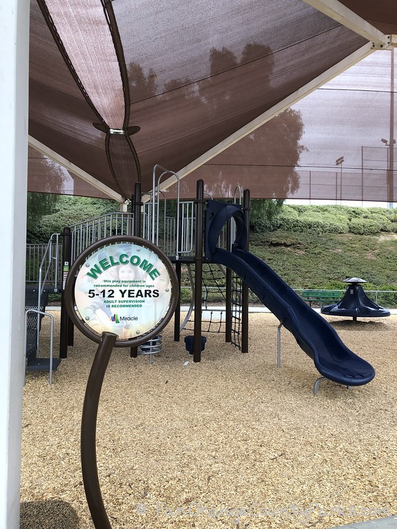 circular sign that says "Welcome: this play equipment is recommended for children ages 5-12 years. Adult supervision is recommended" with a Miracle logo and the playground in the background