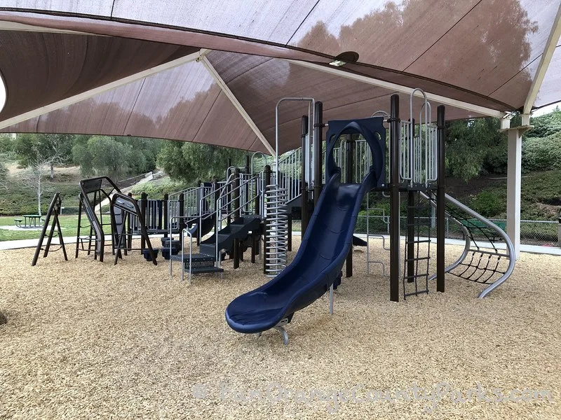 main playground with a navy blue slide and shiny metal corkscrew and stairs on a bark play surface under a brown shade cover