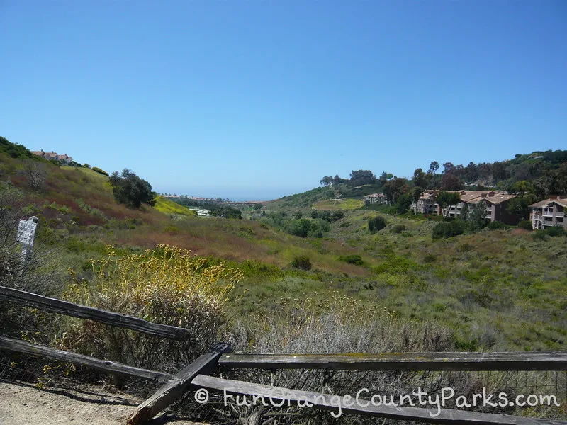 broken wooden fence with a canyon that has bushes and trees with ocean in distance and apartment buildings on the right