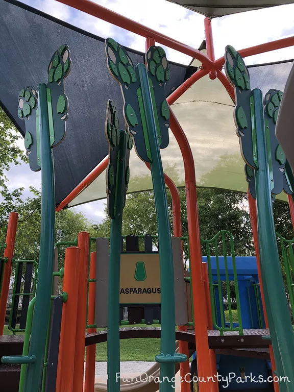 a beige sign with black block writing reads "asparagus" with what appears to be large stalks of asparagus growing up towards the shade sail on the playground (actually climbers for kids)