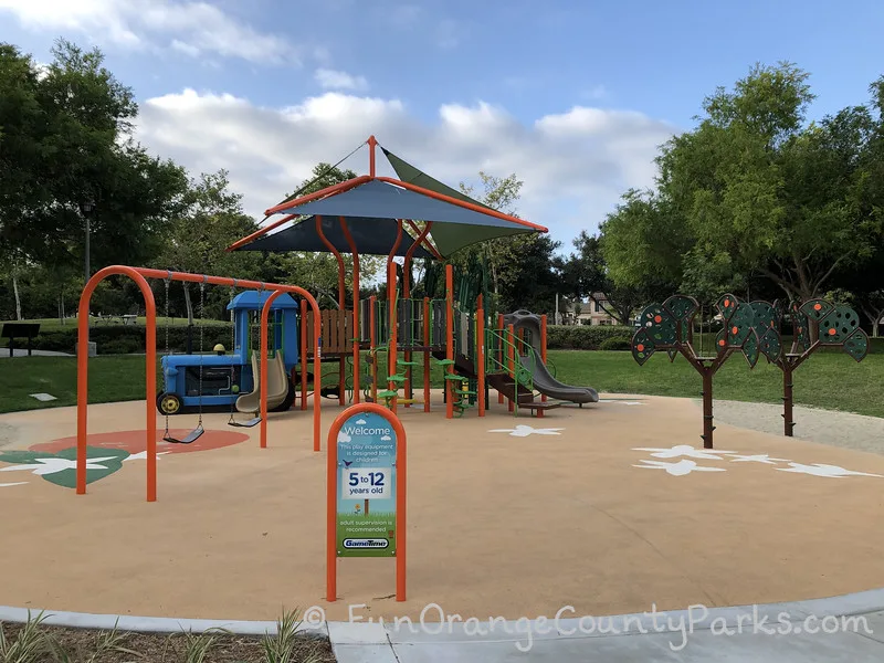 Plaza Park in Irvine 5-12 year old play area on a recycled rubber surface decorated with orange and orange blossoms with a swing set on the left and 2 orange tree climbers on the right. Central is play structure with blue tractor and shade sail.