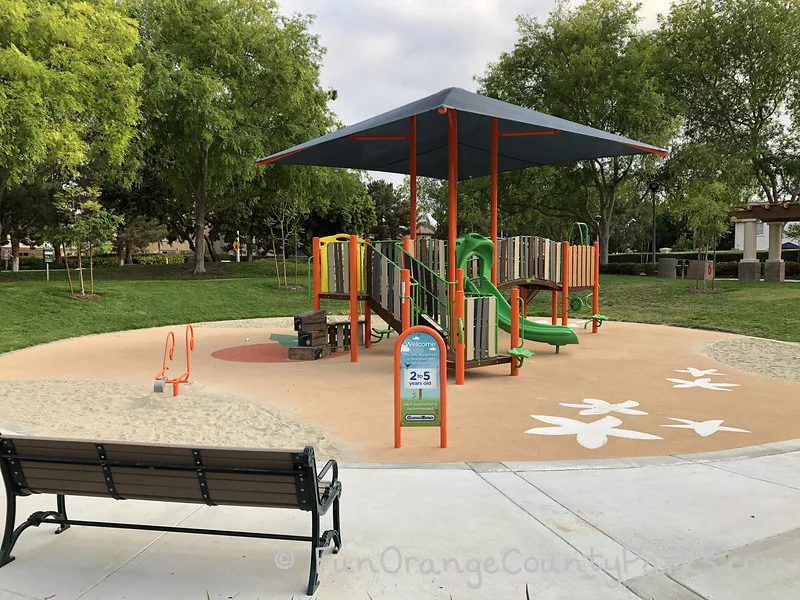 Plaza Park 2-5 year old play structure with view from the back of a bench towards the smaller play equipment with sand areas along the edges, an orange digger/excavator, and green slide under a blue shade sail.