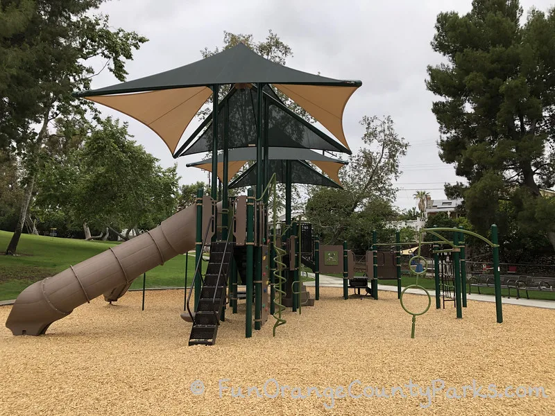 large playground with tan and green shade sails over the play structure on a bark surface with surrounding trees