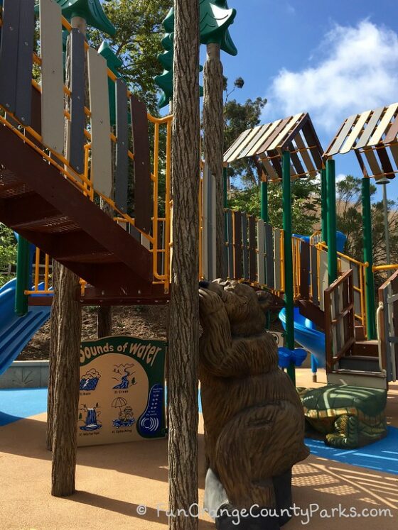 what looks like a lifesize wooden carving of a bear climbing a tree that's actually part of the playground feature of a forest-themed playground at crown valley park in laguna niguel 