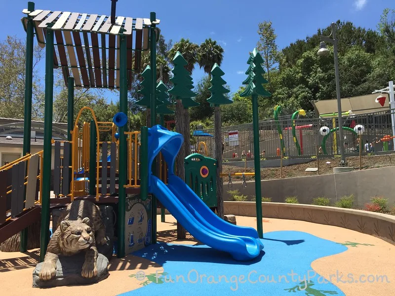 brilliant blue slide coming off small playground with what looks like pine trees and a life-size bobcat playground feature crawling from underneath the equipment -- crown valley park sprayground visible in background raised above the playground