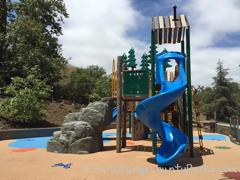 bright blue twisty slides and more rock structures to climb with another view of the playground which shows the recycled rubber surface and blue skies with clouds