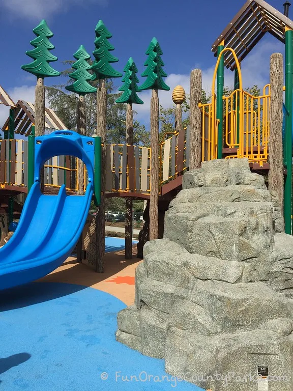 Crown Valley Park playground looks like a real forest with poles textured like pine trees and rocky outcroppings to climb alongside bright blue slides