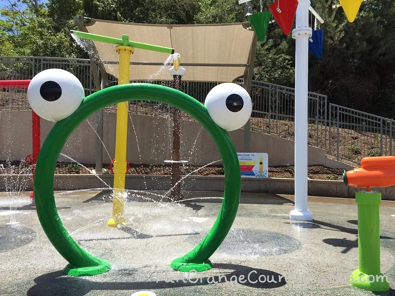 crown valley park laguna niguel splash pad with a bright metal green arch with bulgy eyes on top to help make the sprayer look like a frog with other water features partially visible
