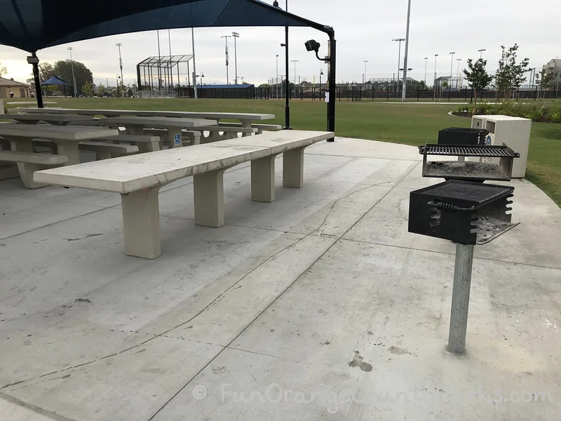 picnic area with the baseball diamond in the background and three barbecue grills in the foreground with a concrete table