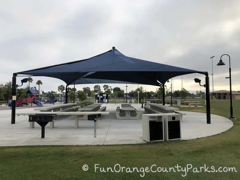 large blue shade structure over concrete picnic tables with grills and trash receptacles in the foreground