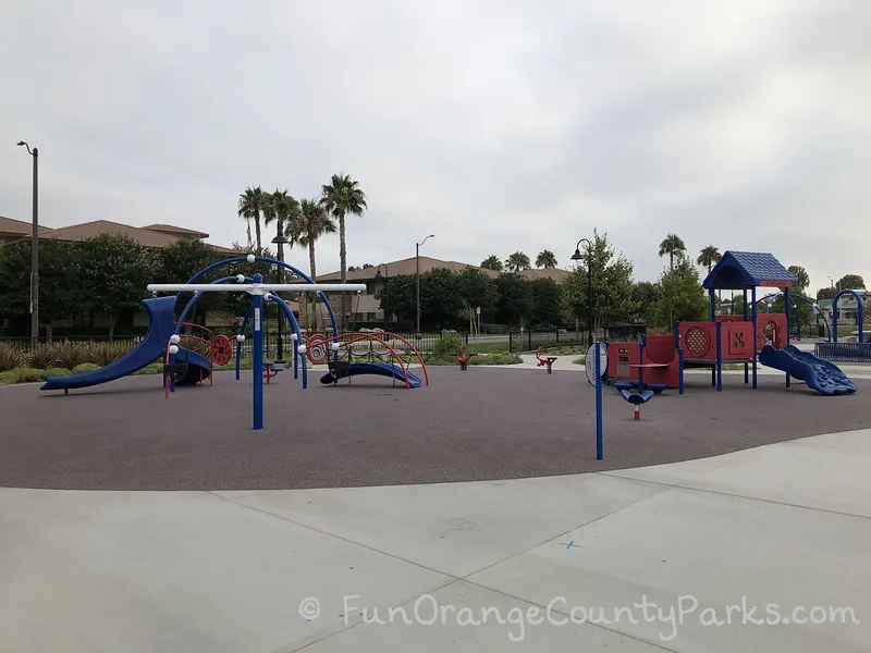 preschool play area at Veterans Sports Park at Tustin Legacy with baby swings, two small play structures, a spinner, and two ride-on toys on a flat recycled rubber surface