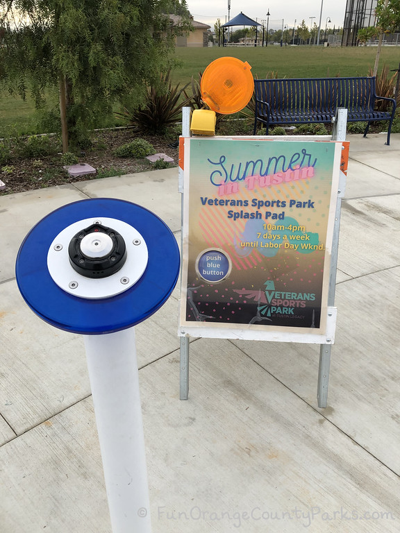a pedestal with a blue button about the size of an adult's palm with a sign that says "Summer in Tustin: Veterans Sports Park Splash Pad 10am-4pm 7 days a week until Labor Day Wknd" and a blue circle on the sign that says "push the blue button" along with the Veterans Sports Park logo