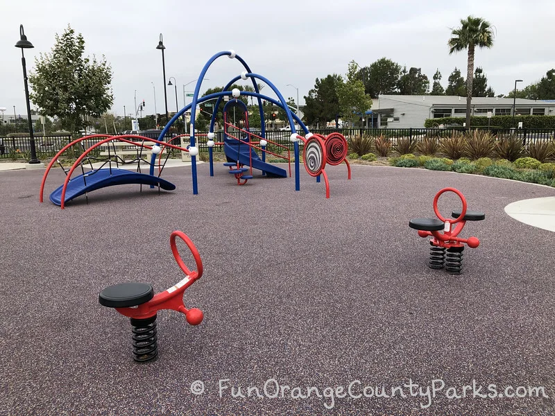 two small ride-on red spring play toys on rose colored recycled rubber surface with blue and red play structure in the background