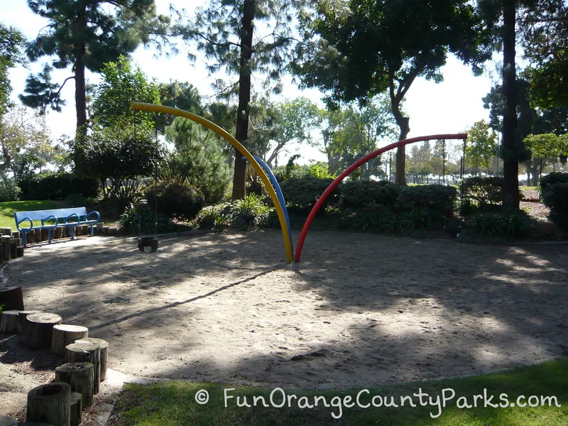 sandy area at Atlantis Play Center in Garden Grove with what looks like 3 bent poles sprouting from the ground with 3 baby swings hanging from the tips of the blue, yellow, and red poles