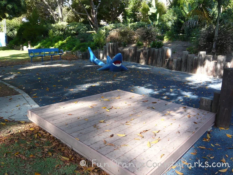small raised wooden stage in a shady park setting with a blue shark for climbing and a blue bench