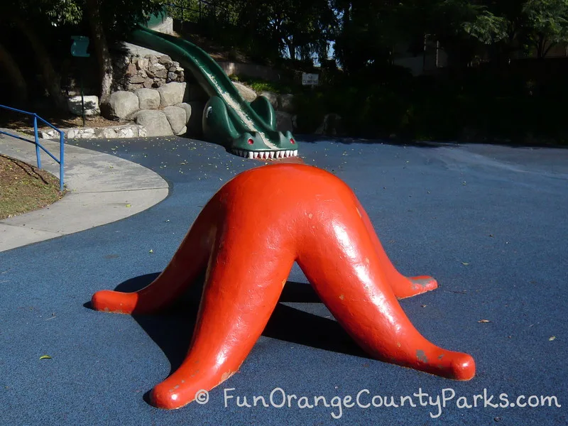 famous green danny the sea dragon slide at Atlantis Play Center with the end of the slide that looks like dragon teeth and a head biting into the blue play surface and the tail stretching up into the trees. Large orange sea star in the foreground. All large enough for children to climb.