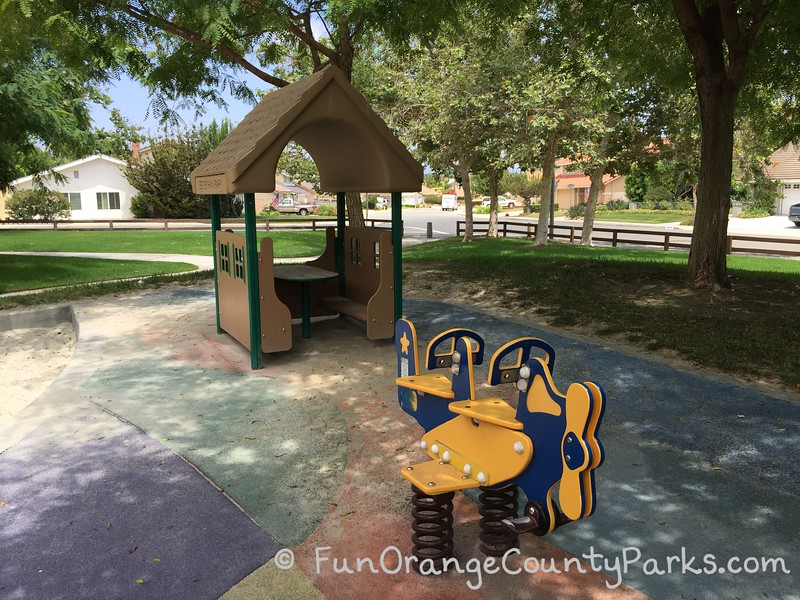 View of beige playground clubhouse at Beckenham Park in Laguna Hills with a bright blue and yellow airplane spring toy with space for two riders on colorful play surface surrounded by green in a neighborhood.