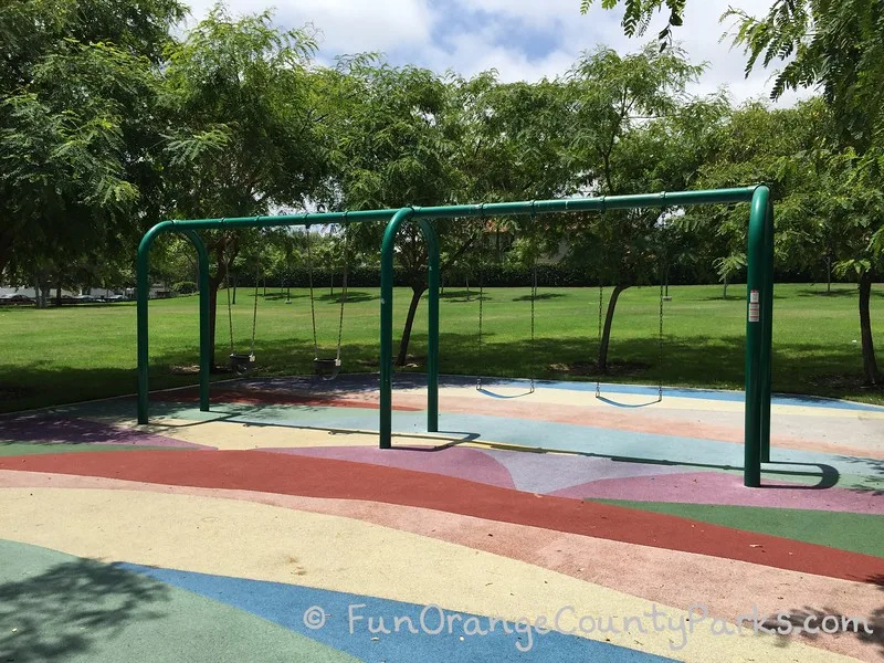 rainbow-colored playground surfacing below swingset with 2 bench swings and 2 baby swings. large grassy green area with shade trees in the background at Beckenham Park in Laguna Hills