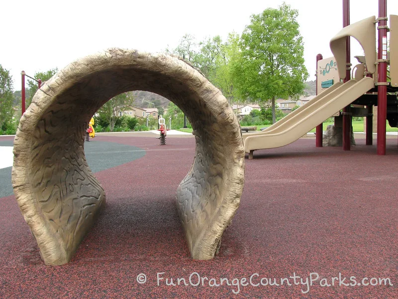 View of Wildcatters Park playground through a log tunnel play structure with slides on the left and spring ride-on toys