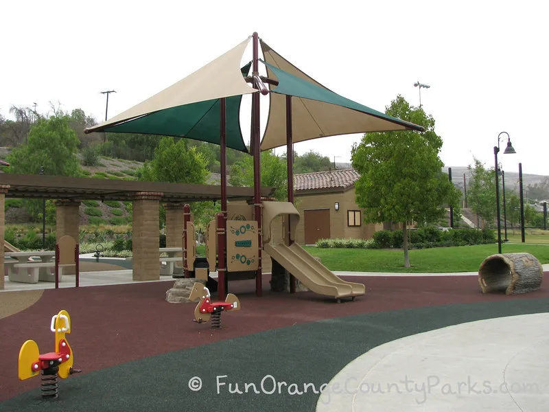 Another playground view of the ride on toys and small playground with a double slide and nearby log tunnel. Covered picnic area behind.