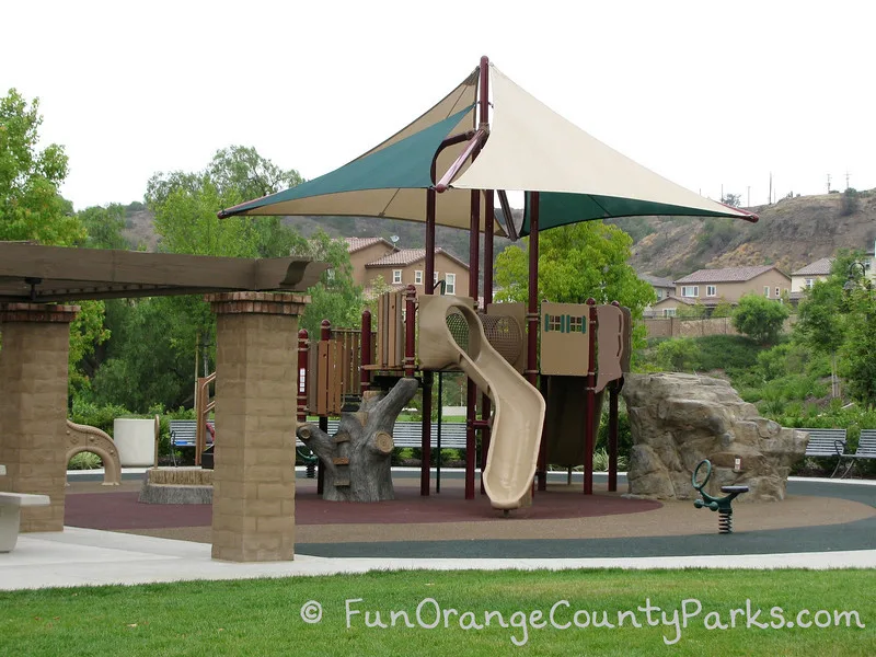 Wildcatters Park in Brea playground with rock for climbing and tree trunk ladder on recycled rubber playground surface. Neighborhood houses in the background.