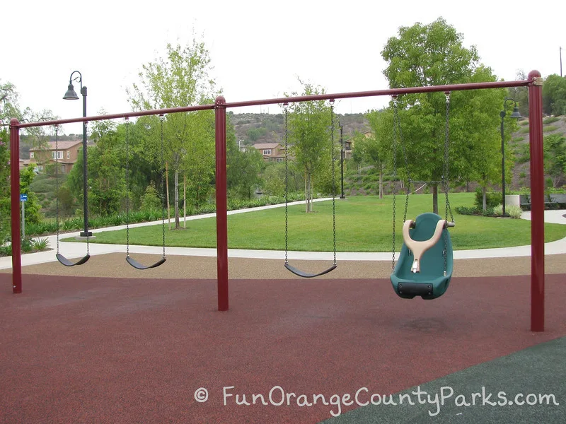 3 bench swings and 1 accessible over red recycled rubber with park setting in the background with lawn and trees